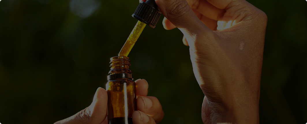 Hands holding an amber CBD oil dropper bottle outdoors against a dark green background